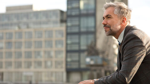 Mann steht auf Balkon in Anzugsjacke. Im Hintergrund befindet sich ein Gebäudekomplex.