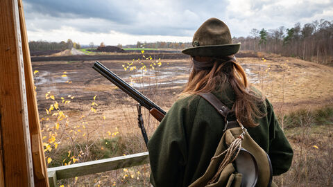 Frau steht mit Rücken zum Fotograf auf einem Hochsitz in Jagdmontur mit Gewehr