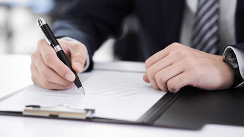 Ein Mann unterschreibt mit einem Kugelschreiber ein Dokument auf einem Klemmbrett. Zu sehen ist nur der Oberkörper und die Hände.