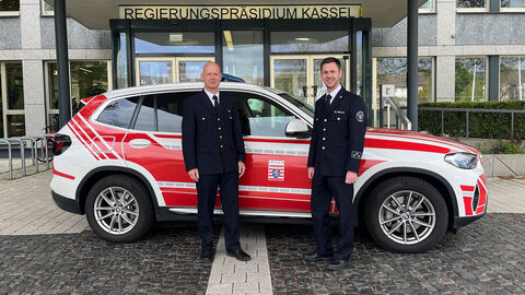Zwei Kollegen vor einem rot/weißen Auto am Haupteingang des RP Kassel.