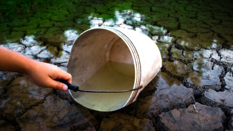 Eine Hand schöpft mit einem alten Eimer Wasser aus einem ausgetrockneten Flussbett