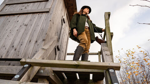 Frau in Jägerbekleidung steht auf einem Hochsitz und schaut in die Ferne