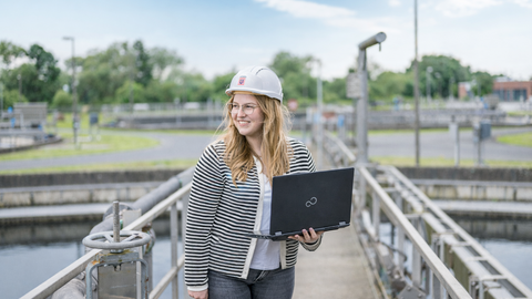 Eine Frau mit Schutzhelm steht auf einem Steg eines Klärbeckens, hält einen Laptop in der Hand und schaut nach links in die Ferne. 